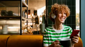 Eine Frau mit Kopfhörern sitzt in einem Café und lächelt mit dem Smartphone in der Hand