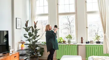 Eine Mutter schaut ihr Kind in den Armen an. Sie steht im Wohnzimmer.