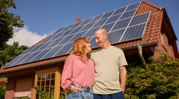 Ein lächelndes Pärchen steht vor ihrem Haus und das Dach ist mit einer PV-Anlage ausgestattet.