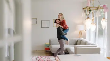 Ein Pärchen freut sich in einer hellen Wohnung vor einem Sofa, der Mann hebt die Frau hoch