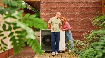 Ein lächelndes Pärchen steht bei einer Wärmepumpe vor einem Backsteinhaus.