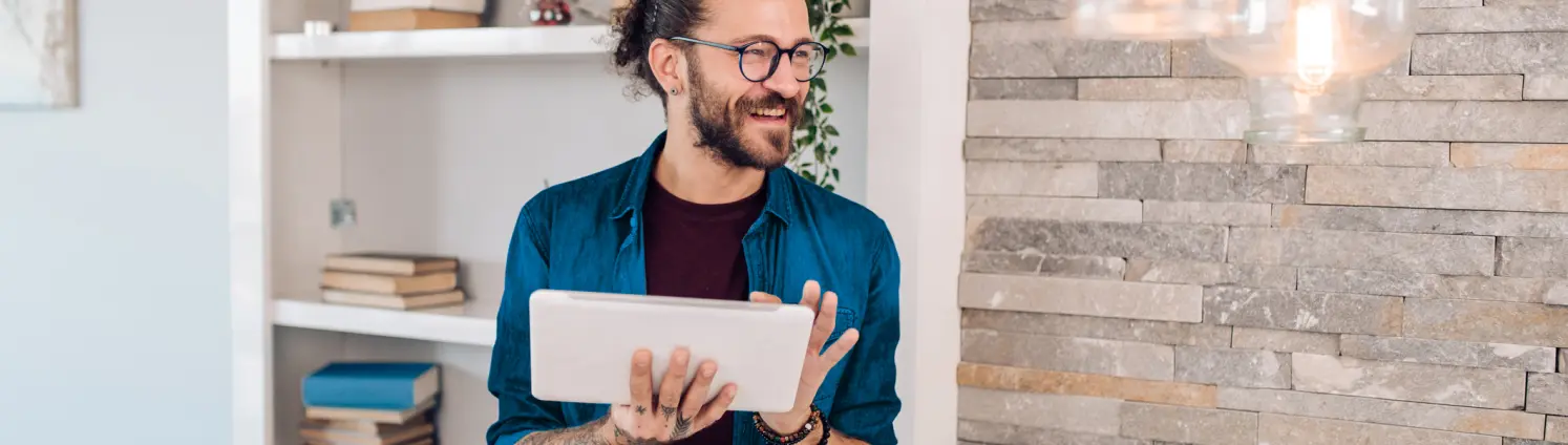 Ein Mann in blauem Hemd steht lächelnd in seiner modernen Wohnung und hält ein Tablet in seiner Hand