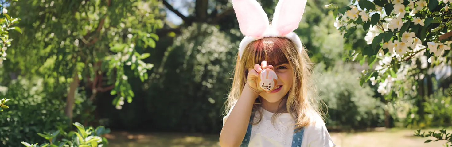 Ein junges Mädchen trägt rosafarbene Hasenohren. Sie hält sich ein Osterei vor das Gesicht, auf das ein Osterhase gemalt ist.