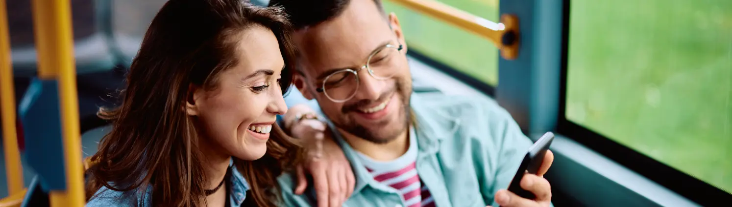 Lachendes Pärchen sitzt im Bus und schaut gemeinsam auf ein Handy