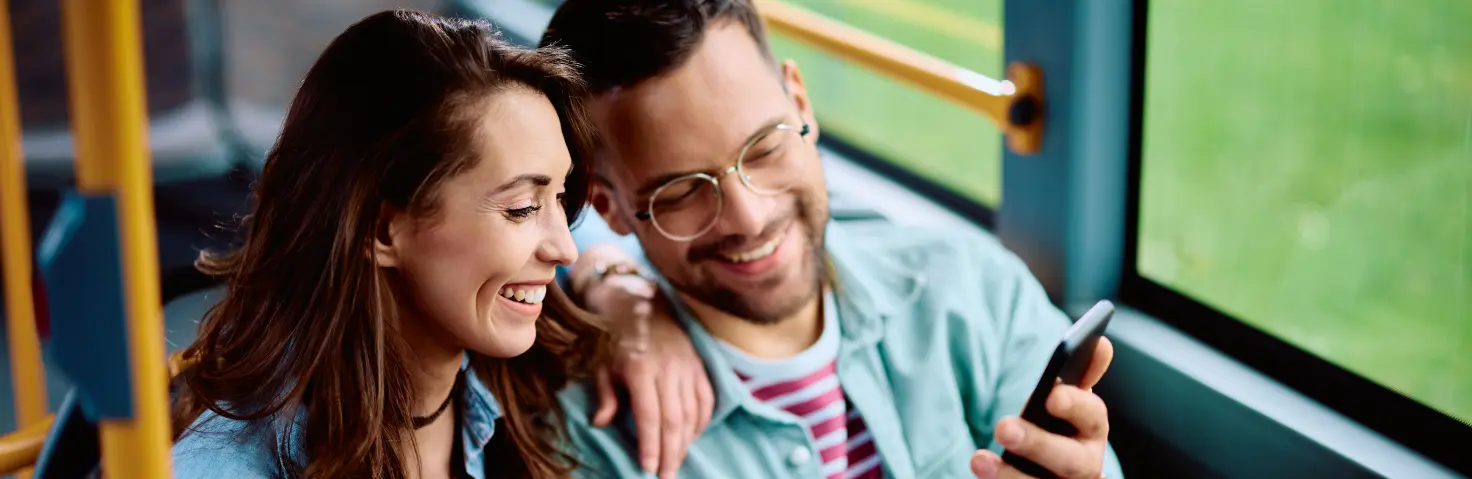 Lachendes Pärchen sitzt im Bus und schaut gemeinsam auf ein Handy