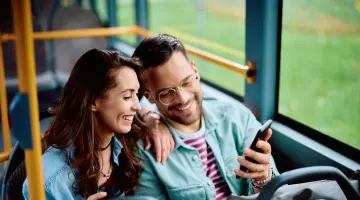 Lachendes Pärchen sitzt im Bus und schaut gemeinsam auf ein Handy