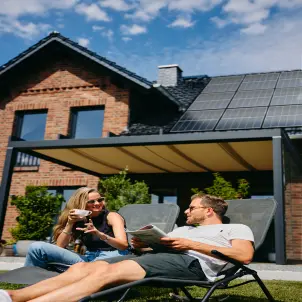 Mann und Frau sitzen im Liegestuhl vor einem Haus mit Photovoltaik-Anlage und lesen eine Zeitschrift und trinken Kaffee