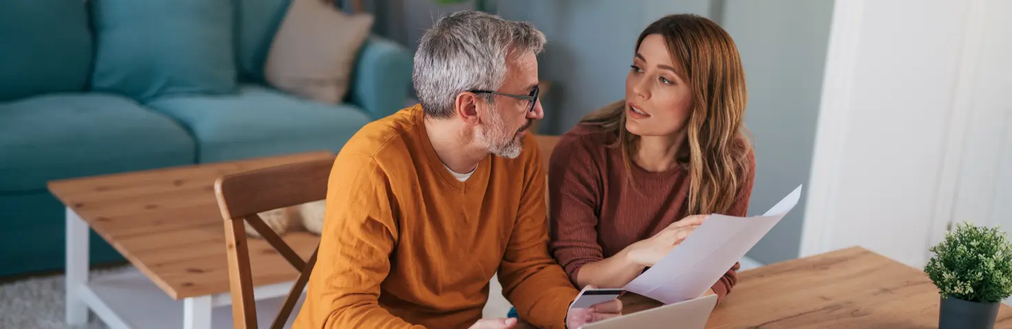 Eine Frau und ein Mann sitzen an einem Tisch und besprechen sich vor einen Laptop und Papieren
