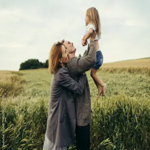 Sommertag: Zwei Erwachsene heben ein Kind vor einem grünen Getreidefeld in die Höhe.