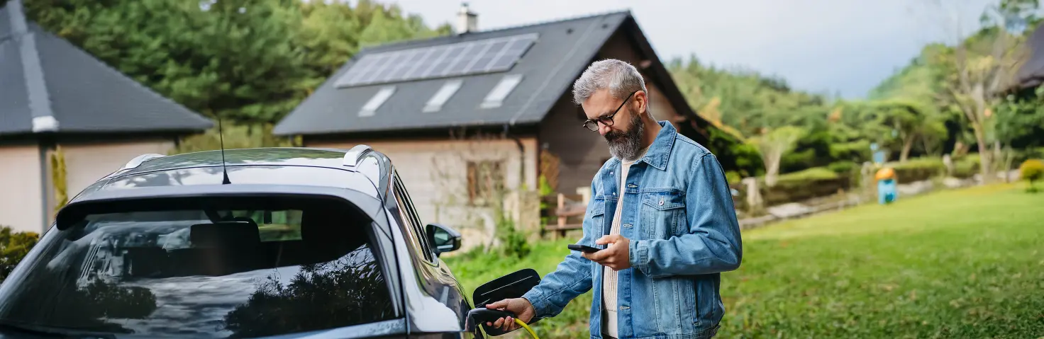 Ein Mann tankt sein E-Auto und guckt nebenbei auf sein Handy. Im Hintergrund ist ein haus mit Photovoltaikanlage.