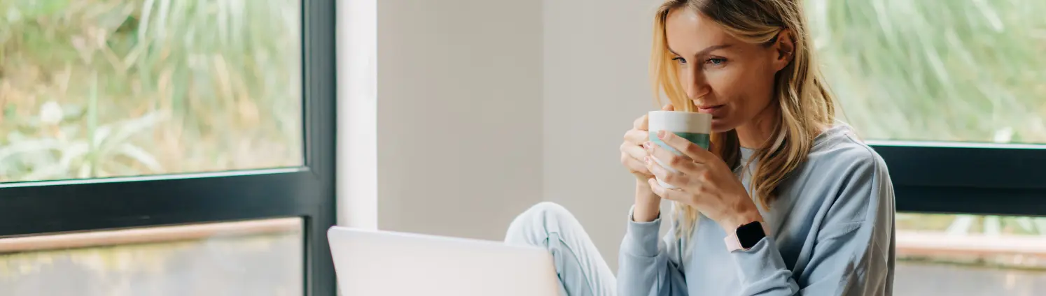 Frau sitzt in ihrer Wohnung und schaut auf ihren Laptopbildschirm mit einer Tasse Kaffee in der Hand