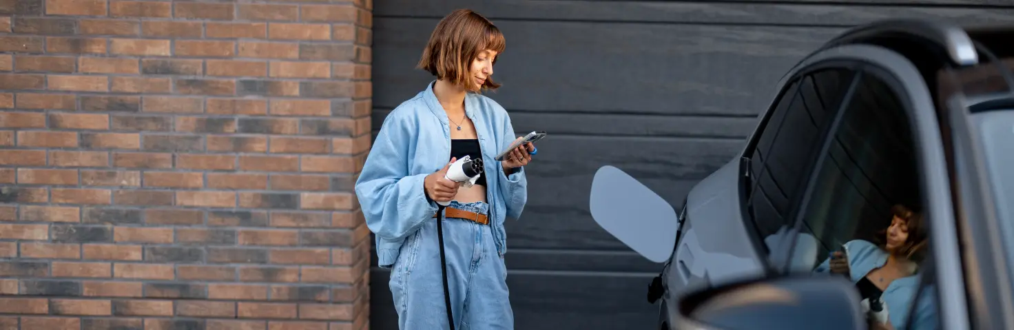 Eine Frau steht mit ihrem Handy und einem Ladestecker in den Händen neben ihrem E-Auto