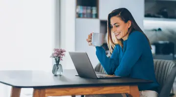 Eine Frau mit Tasse in der Hand schaut lächelnd auf ihren Laptop