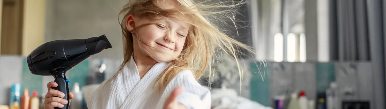 Ein Mädchen föhnt lächelnd ihre blonden, langen Haare und zeigt Daumen hoch in die Kamera.