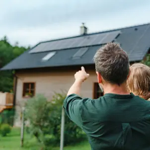 Ein Mann von hinten, trägt seine Tochter auf dem Arm und zeigt auf eine Photovoltaik-Anlage auf einem Dach von einem Haus. Bild mit KI erstellt.