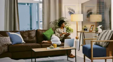 Frau sitzt auf der Couch mit einem Tablet in der Hand und schaut auf die Stehlampe neben ihr. Bild mit KI erstellt.
