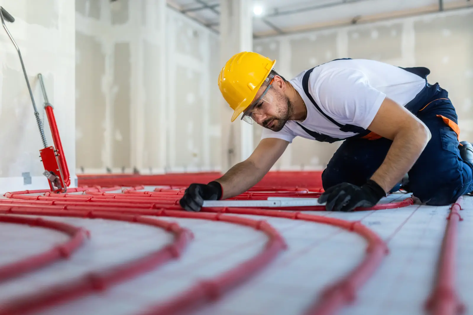 Handwerker mit Helm verlegt die Schläuche einer Fußbodenheizung im Innenraum eines Neubaus. Bild mit KI erstellt.