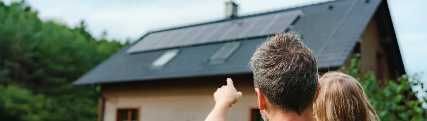 Rückansicht von Papa, der ihr kleines Mädchen in den Armen hält und in ihrem Haus mit installierten Sonnenkollektoren zeigt