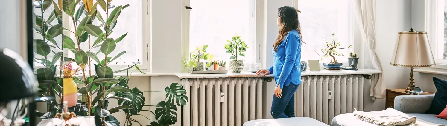 Frau mit mittlerer Erwachsener, die am Heizkörper steht und zu Hause aus dem Fenster schaut – St