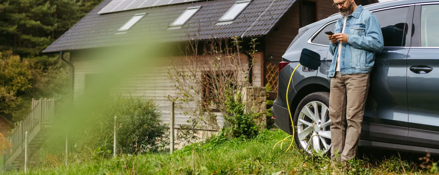 Ein Mann lehnt an seinem Auto welches geladen wird durch die PV-Anlage auf dem Haus hinter ihm