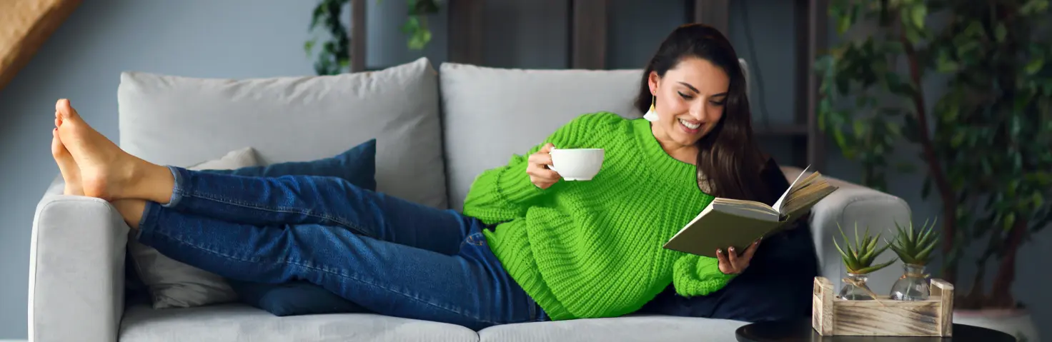 Eine Frau liegt auf ihrem Sofa, trinkt Tee und ließt ein Buch