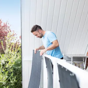 Junger Mann im T-Shirt befestigt ultraleichte Solarmodule am Holzgeländer seines Einfamilienhauses.
