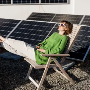 Junge Frau mit Sonnenbrille und grünem Pullover sitzt in einem Sonnenstuhl vor ihrer Photovoltaik-Anlage auf einem Flachdach.