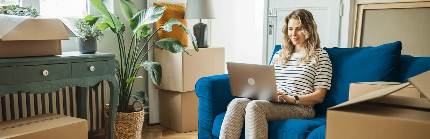 Frau sitzt auf einer blauen Couch und auf ihrem Schoß liegt ihr Laptop, auf dem sie schreibt.