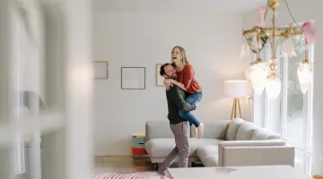 Ein Mann hebt seine Frau hoch, beide freuen sich in ihrer hellen Wohnung