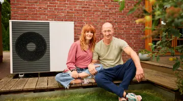 Eine Frau und ein Mann sitzen draußen vor einer Wärmepumpe auf ihrer Terasse