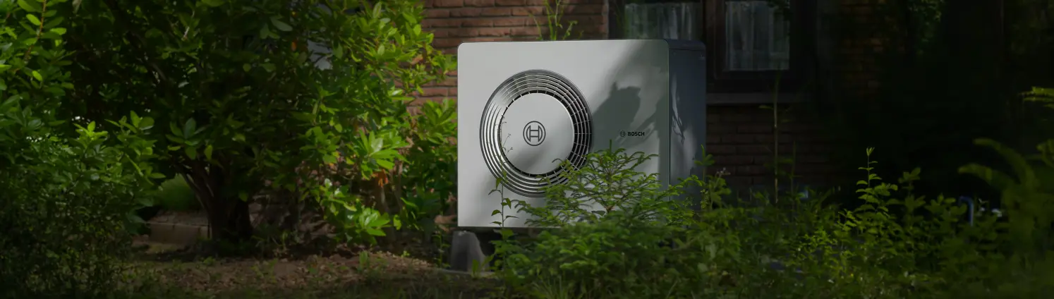 Eine Wärmepumpe steht zwischen Sträuchern im Garten vor einem Haus. Bild mit KI erstellt.