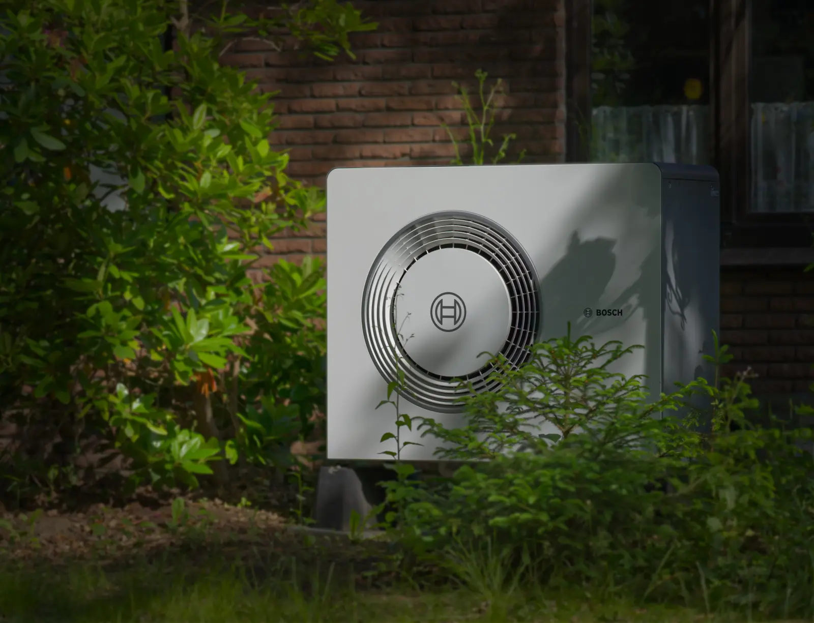 Eine Wärmepumpe steht zwischen Sträuchern im Garten vor einem Haus